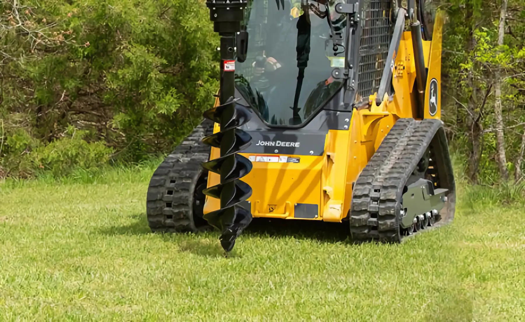 A yellow John Deere skid steer with a large auger attachment is parked on green grass near trees, ready for digging. AdvanceTrac