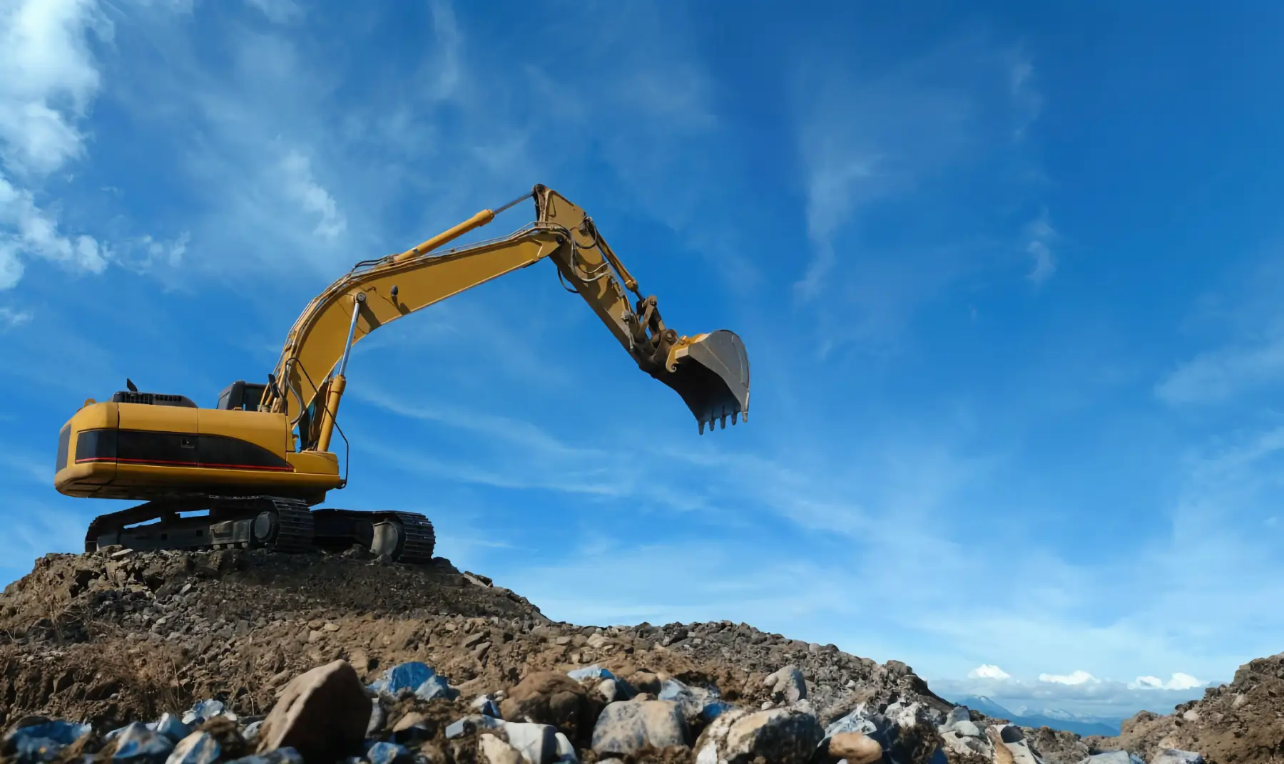 A yellow excavator sits on a pile of dirt and rocks under a bright blue sky with scattered clouds, working at a construction or mining site. AdvanceTrac