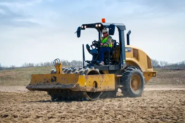 A construction worker operates a yellow soil compactor with spiked rollers, a vital piece of compaction equipment, on a dirt field under a partly cloudy sky. AdvanceTrac