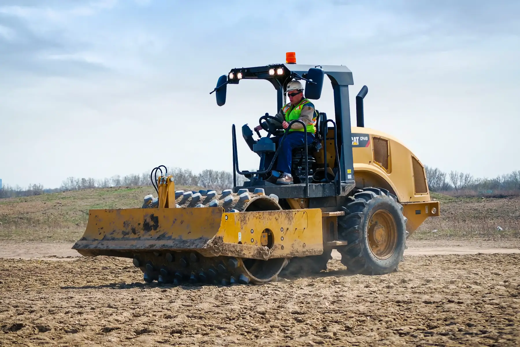 A construction worker operates a yellow soil compactor with spiked rollers, a vital piece of compaction equipment, on a dirt field under a partly cloudy sky. AdvanceTrac