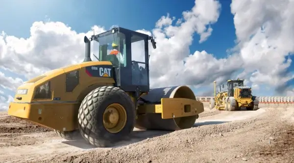 A construction worker operates Compaction Equipment, specifically a CAT compactor, on a dirt road under a blue sky with clouds, with another construction vehicle in the background. AdvanceTrac
