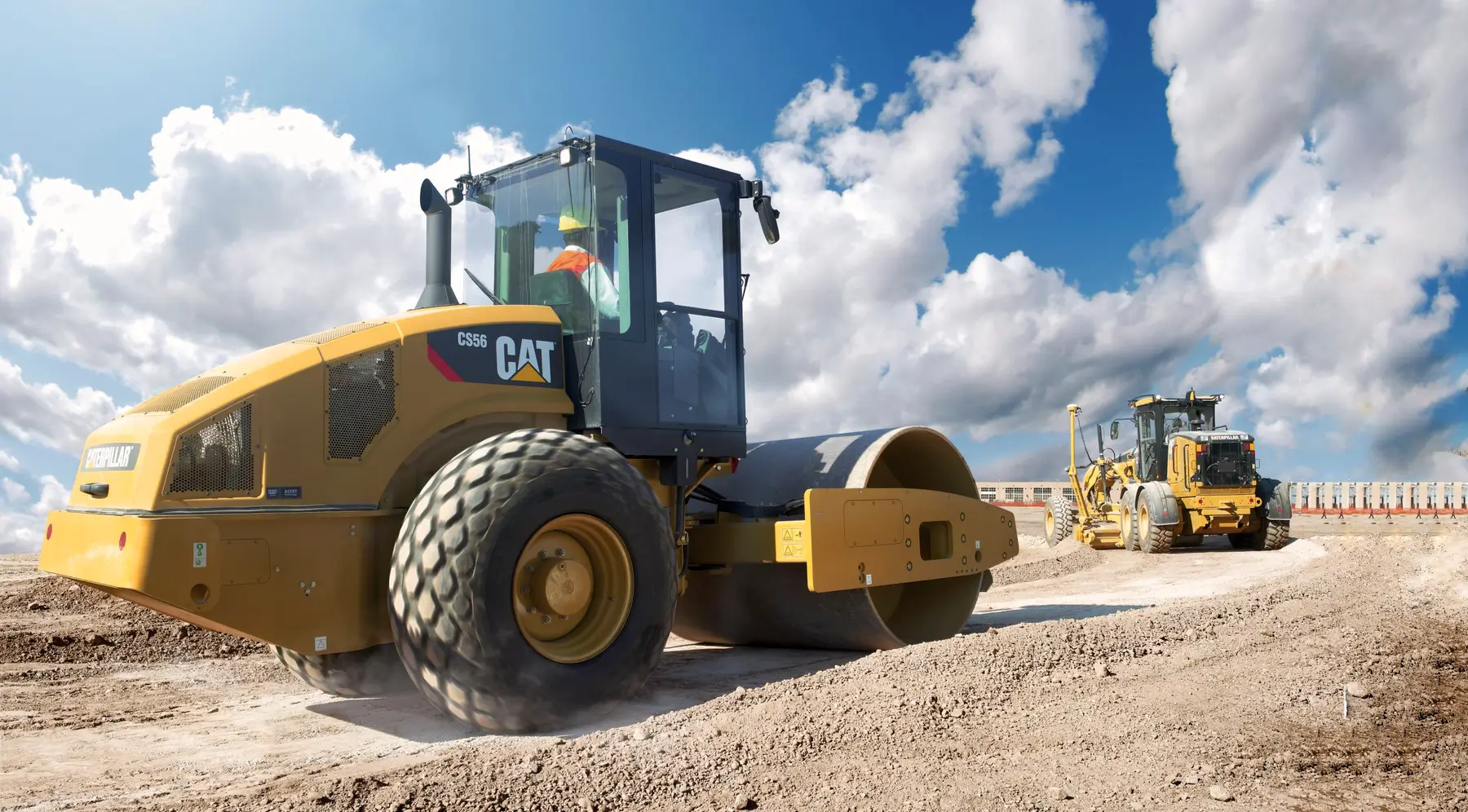 A construction worker operates Compaction Equipment, specifically a CAT compactor, on a dirt road under a blue sky with clouds, with another construction vehicle in the background. AdvanceTrac