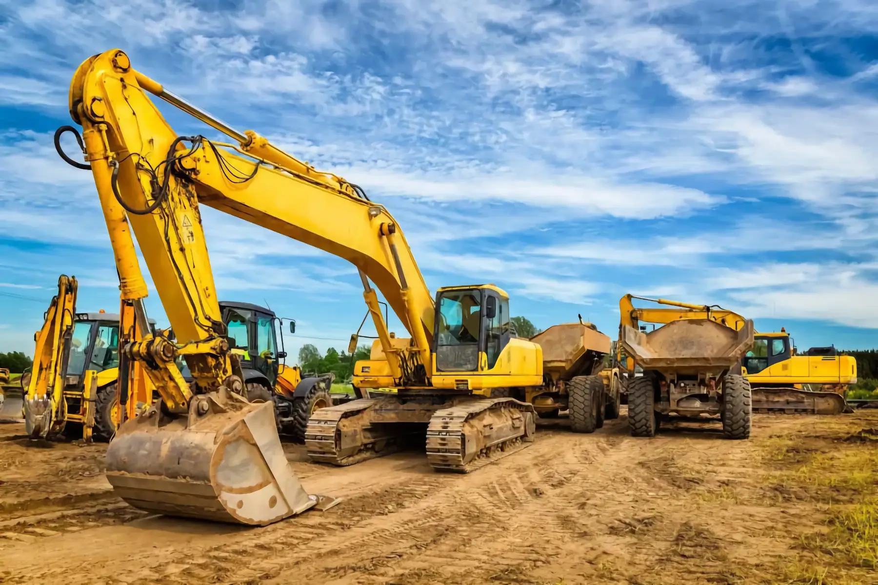 Several large yellow excavators and dump trucks are parked on a dirt construction site under a partly cloudy blue sky. AdvanceTrac