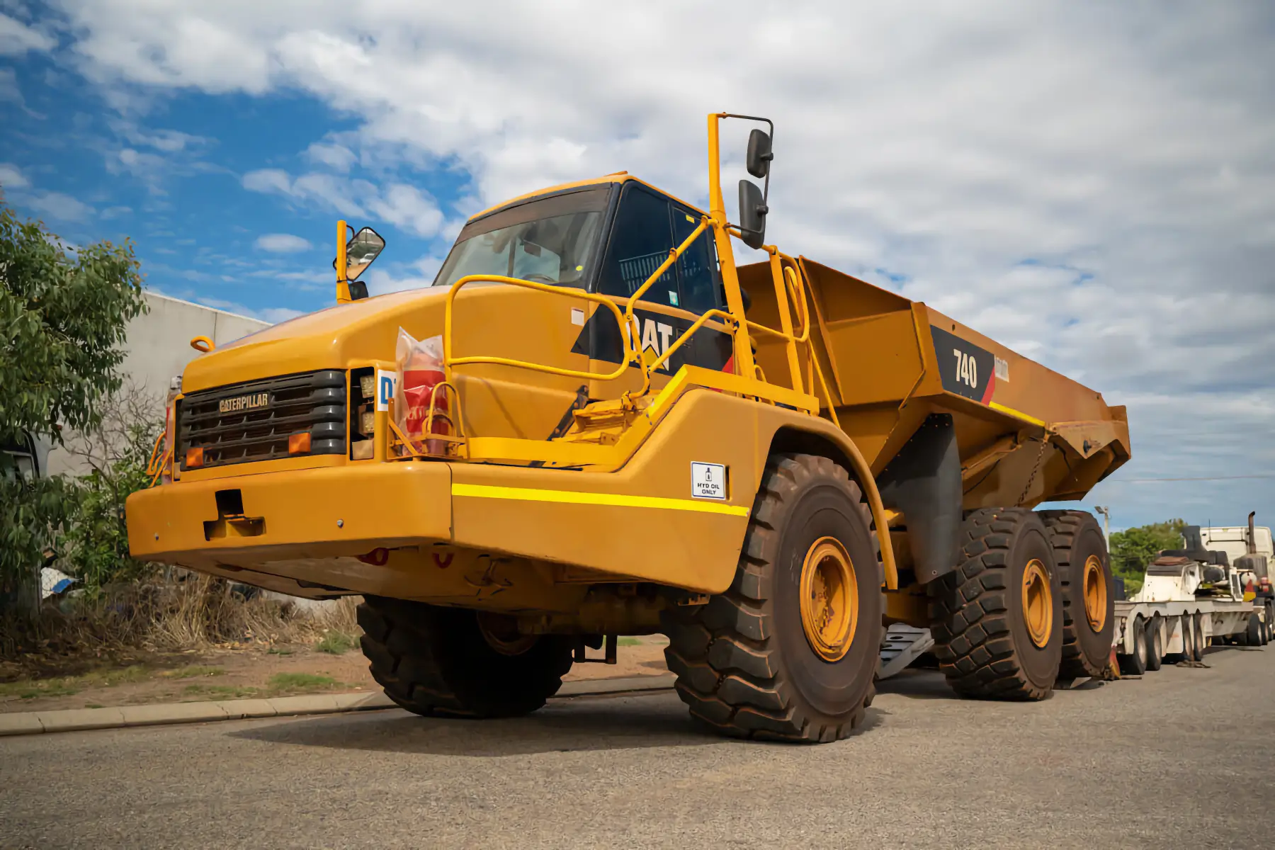 A large yellow Caterpillar 740 articulated dump truck is parked on a paved road under a partly cloudy sky. Trees and a building are visible in the background. AdvanceTrac