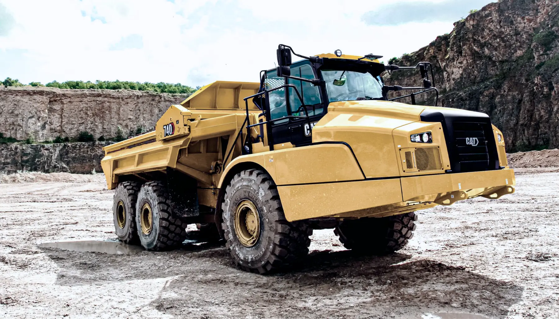 A large yellow articulated off-road dump truck with six wheels is parked at a quarry site, surrounded by rocky terrain and cliffs under a partly cloudy sky. AdvanceTrac