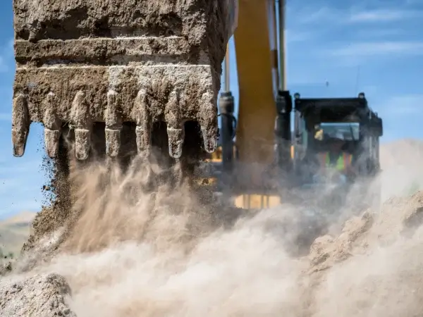 A close-up of an excavator bucket digging into dirt, releasing dust and soil, with a construction worker visible in the blurred cab in the background. AdvanceTrac