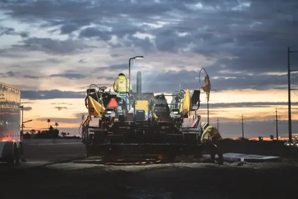 Two construction workers operate heavy machinery on a road at dusk, with streetlights and a dramatic cloudy sky in the background. AdvanceTrac