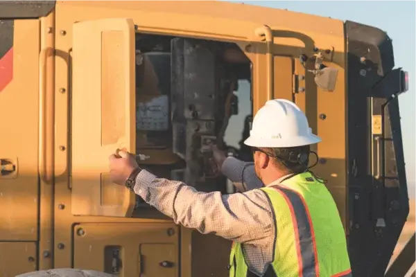 A construction worker wearing a white hard hat and reflective vest opens a panel on a large yellow industrial machine under daylight. AdvanceTrac