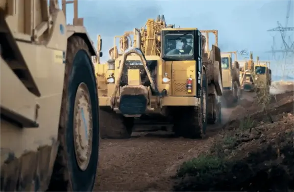 Several large yellow construction vehicles drive on a dirt road at a work site, kicking up dust, with one vehicle prominently in the foreground and others following behind. AdvanceTrac