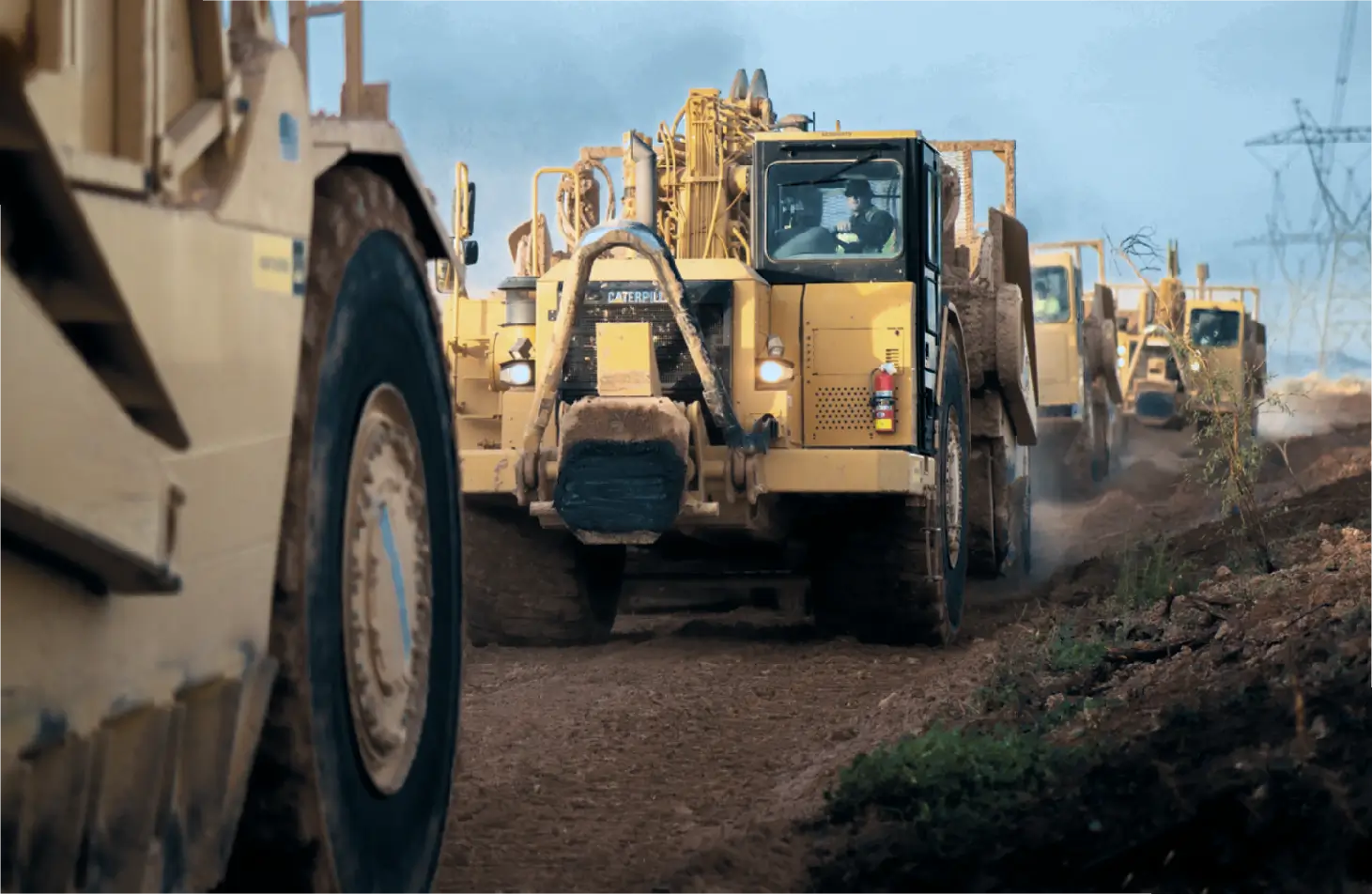 Several large yellow construction vehicles drive on a dirt road at a work site, kicking up dust, with one vehicle prominently in the foreground and others following behind. AdvanceTrac