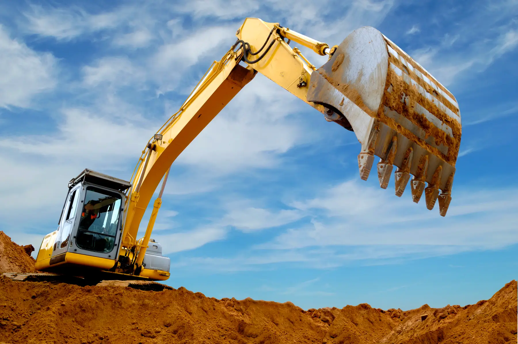 A yellow excavator with a large bucket is digging into a mound of brown dirt under a blue sky with white clouds. AdvanceTrac