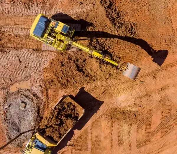Aerial view of a yellow excavator loading dirt into a dump truck on a construction site with reddish-brown soil and tire tracks. AdvanceTrac