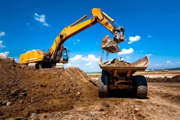 A yellow excavator loads dirt into a dump truck at a construction site under a bright blue sky with scattered clouds. AdvanceTrac