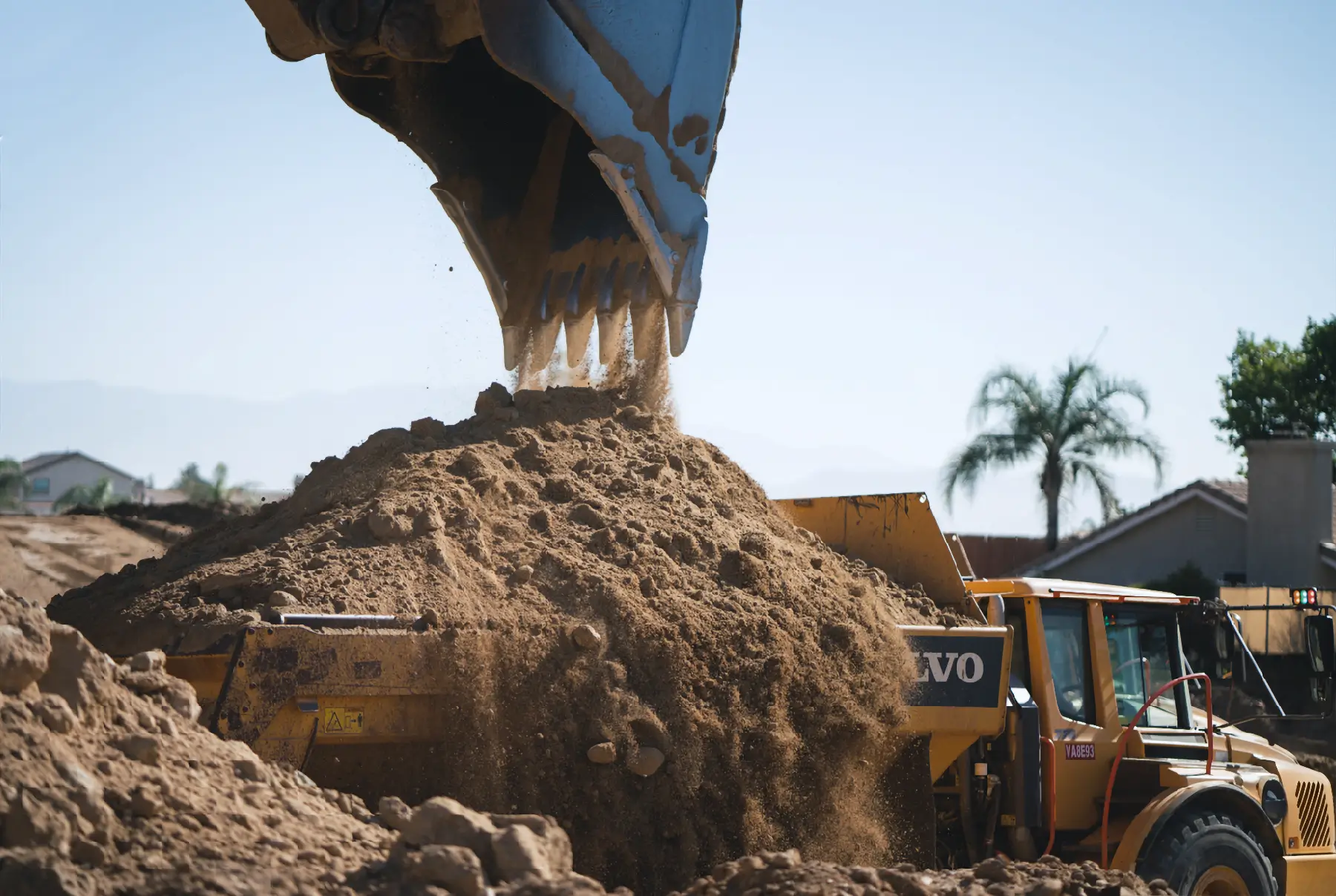 A large excavator dumps a pile of dirt onto a construction site, with a yellow Volvo vehicle nearby and palm trees in the background. AdvanceTrac