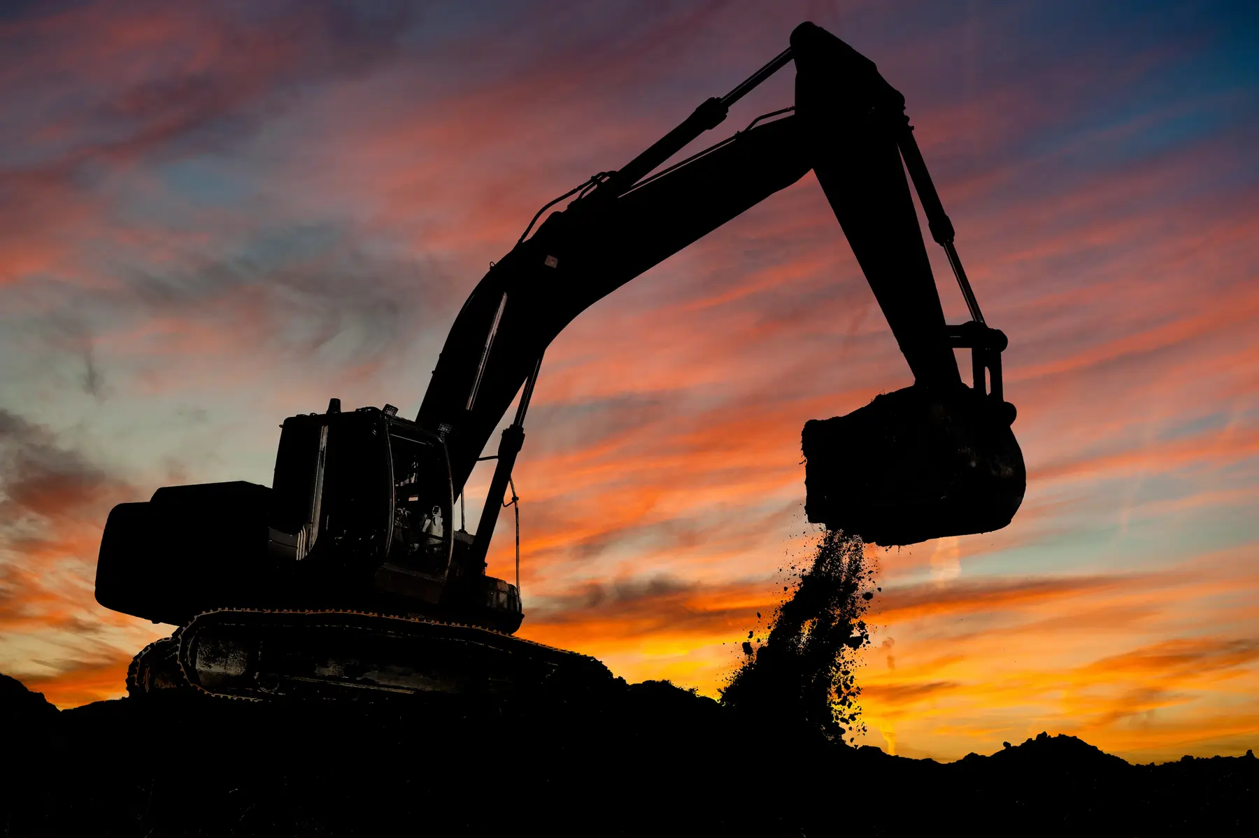 Silhouette of an excavator digging and dumping dirt at a construction site during a vibrant sunset with colorful clouds in the sky. AdvanceTrac