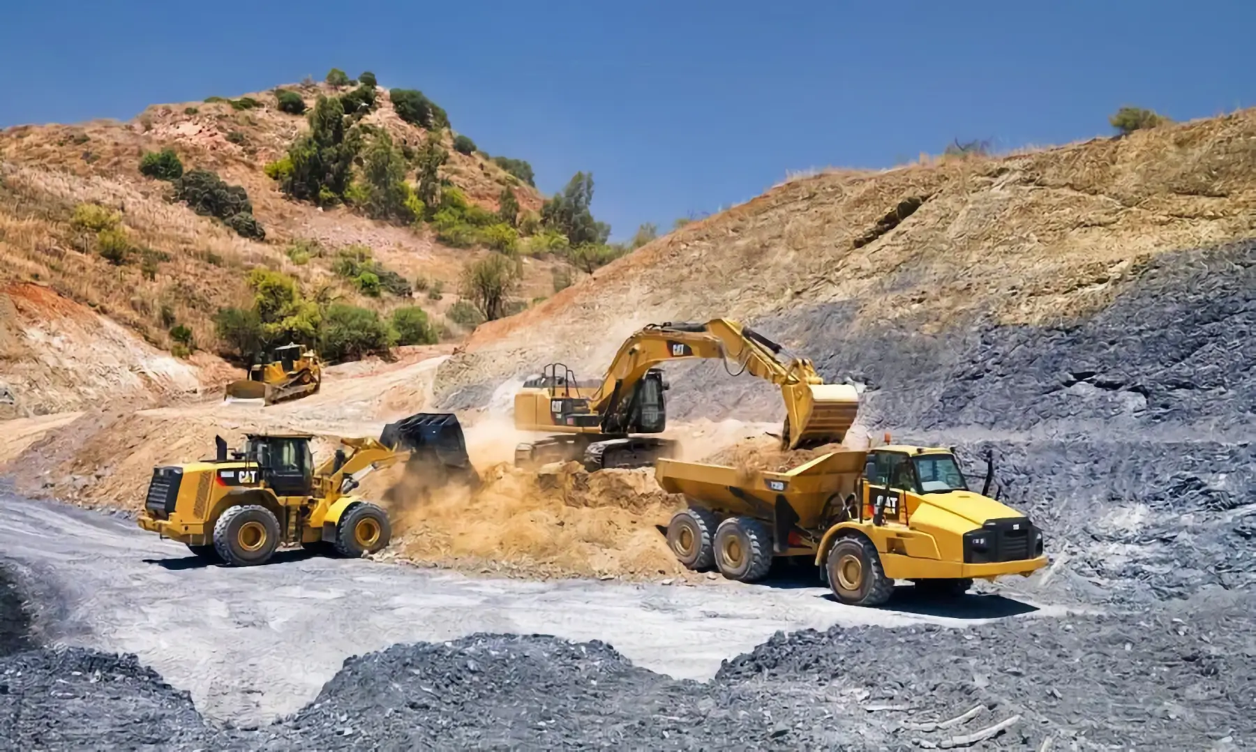 Yellow construction vehicles, including an excavator, dump truck, and loader, work on a dusty, rocky landscape at a mining or construction site under a clear blue sky with hills and trees in the background. AdvanceTrac