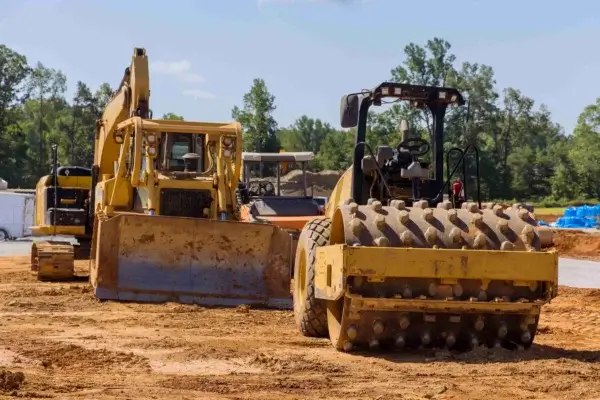 A bulldozer and a compactor sit on a construction site with dirt and trees in the background under a clear sky. AdvanceTrac