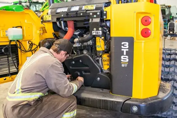 A mechanic in coveralls and a cap works on the engine of a yellow construction vehicle labeled “3TS 8T” inside a garage. AdvanceTrac