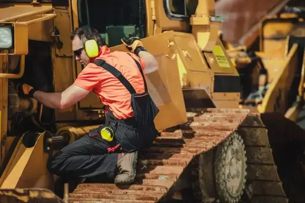 A worker in safety gear and ear protection repairs or inspects a large yellow construction vehicle while kneeling on its metal tracks. AdvanceTrac