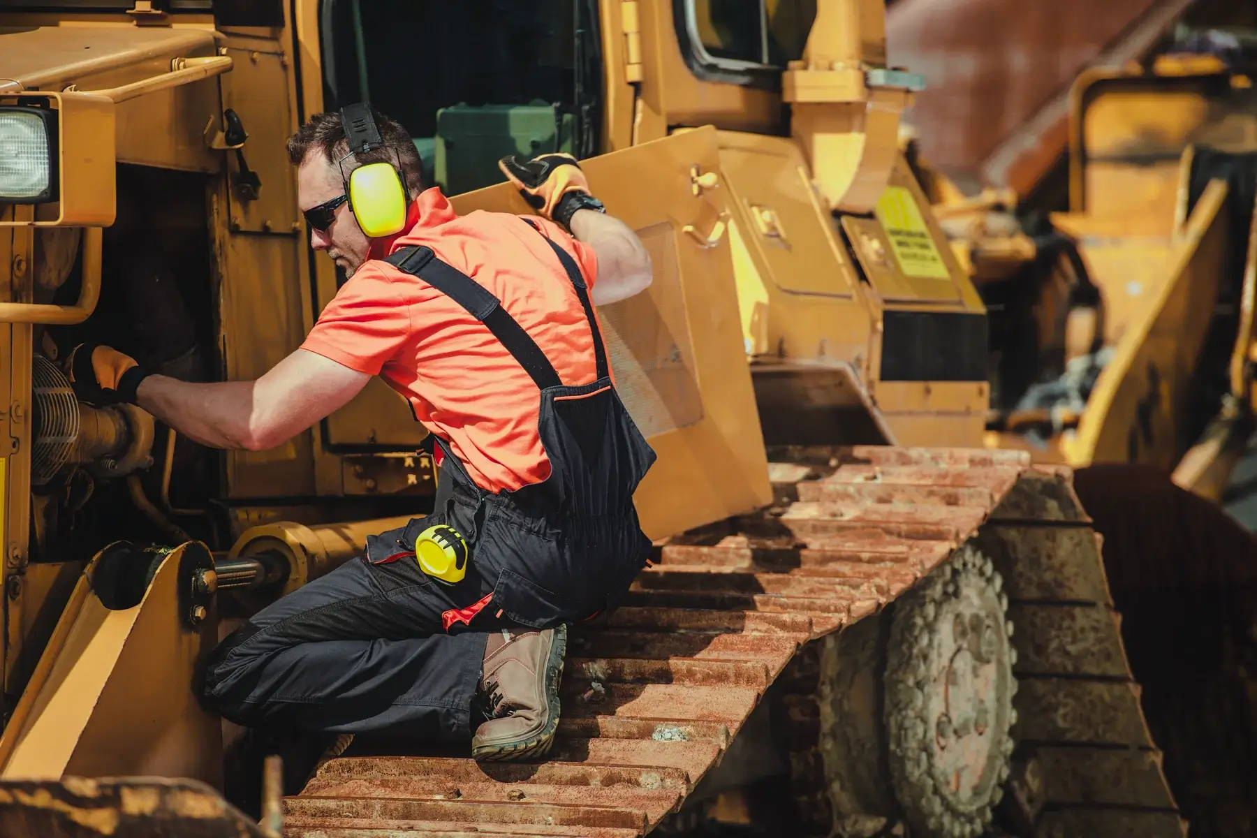 A worker in safety gear and ear protection repairs or inspects a large yellow construction vehicle while kneeling on its metal tracks. AdvanceTrac
