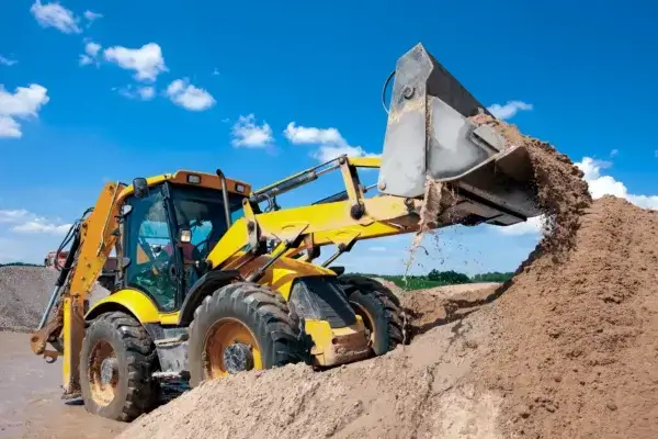 A yellow backhoe loader lifts and dumps sand onto a large pile at a construction site under a bright blue sky with scattered clouds. AdvanceTrac