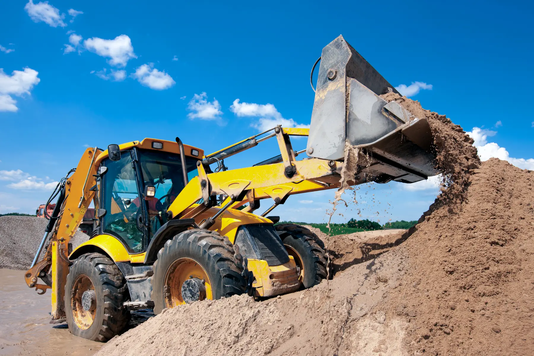 A yellow backhoe loader lifts and dumps sand onto a large pile at a construction site under a bright blue sky with scattered clouds. AdvanceTrac