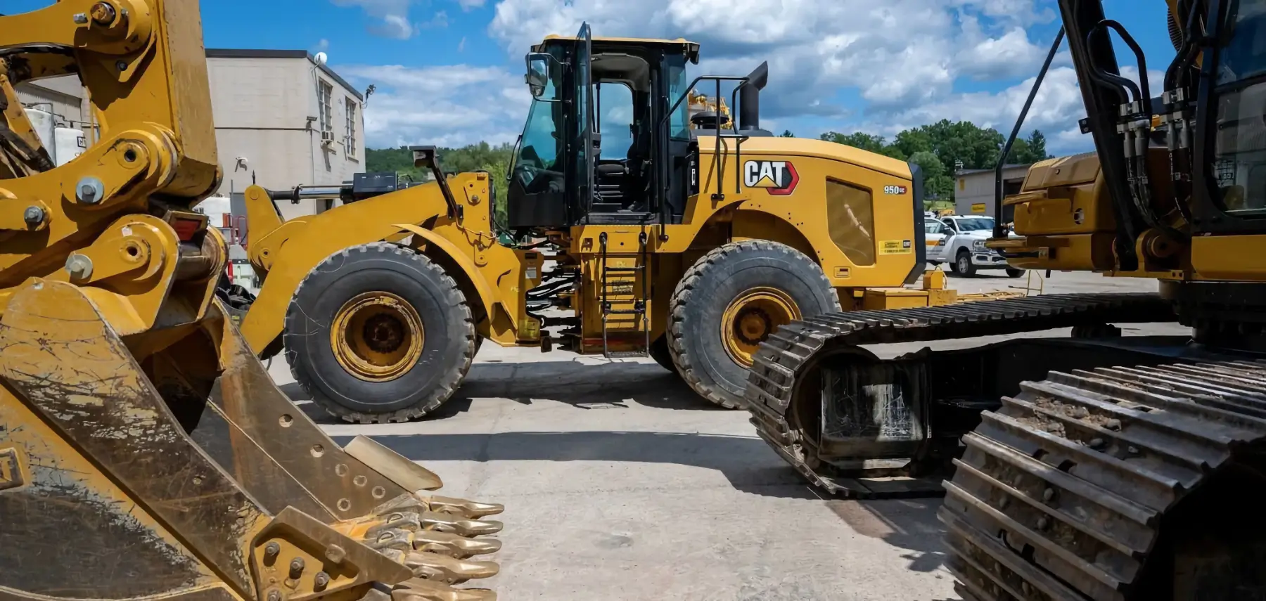 A yellow Caterpillar wheel loader is parked outside, surrounded by other construction equipment, including a tracked excavator and a large bucket, on a sunny day with scattered clouds. AdvanceTrac