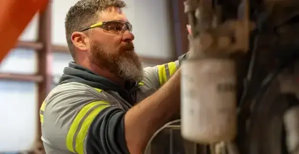 A bearded man wearing safety glasses and a gray shirt with yellow reflective stripes works on machinery in an industrial setting. AdvanceTrac