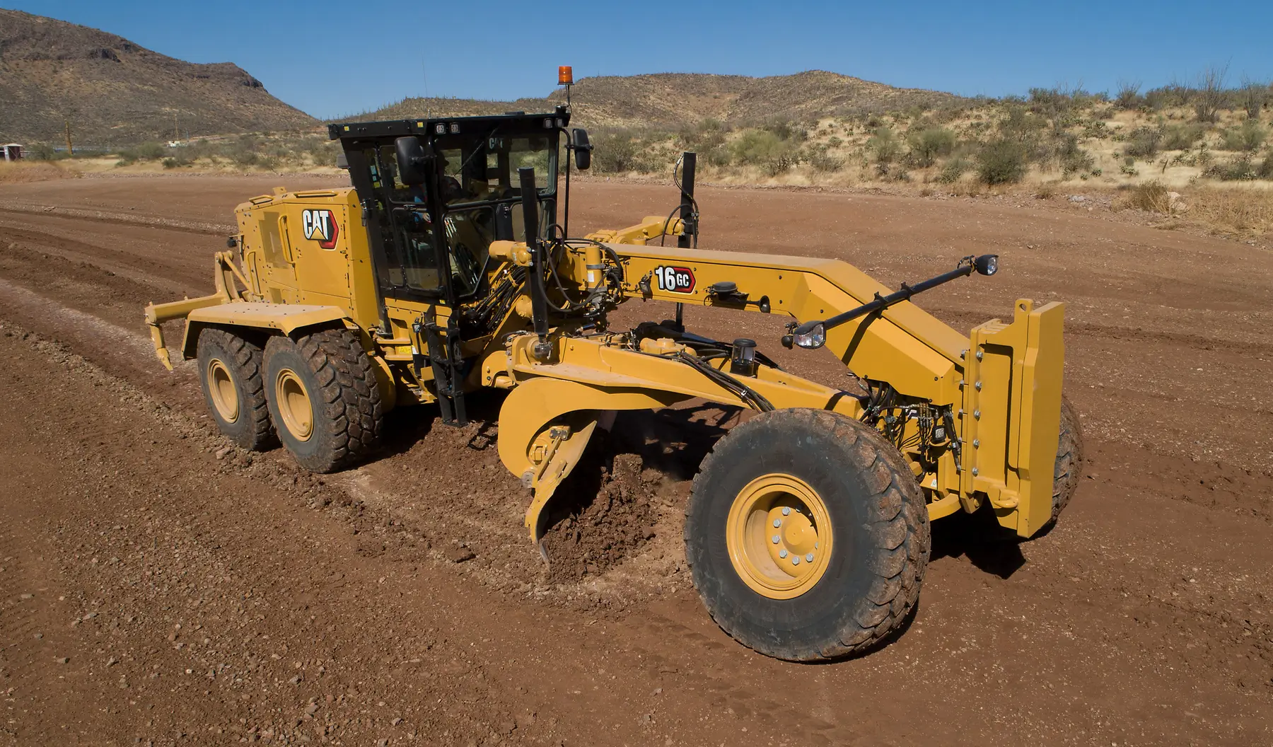 A yellow Caterpillar motor grader levels a dirt road in a dry, desert landscape with hills and sparse vegetation under a clear blue sky, showcasing the reliability and precision of Motor Graders in tough environments. AdvanceTrac
