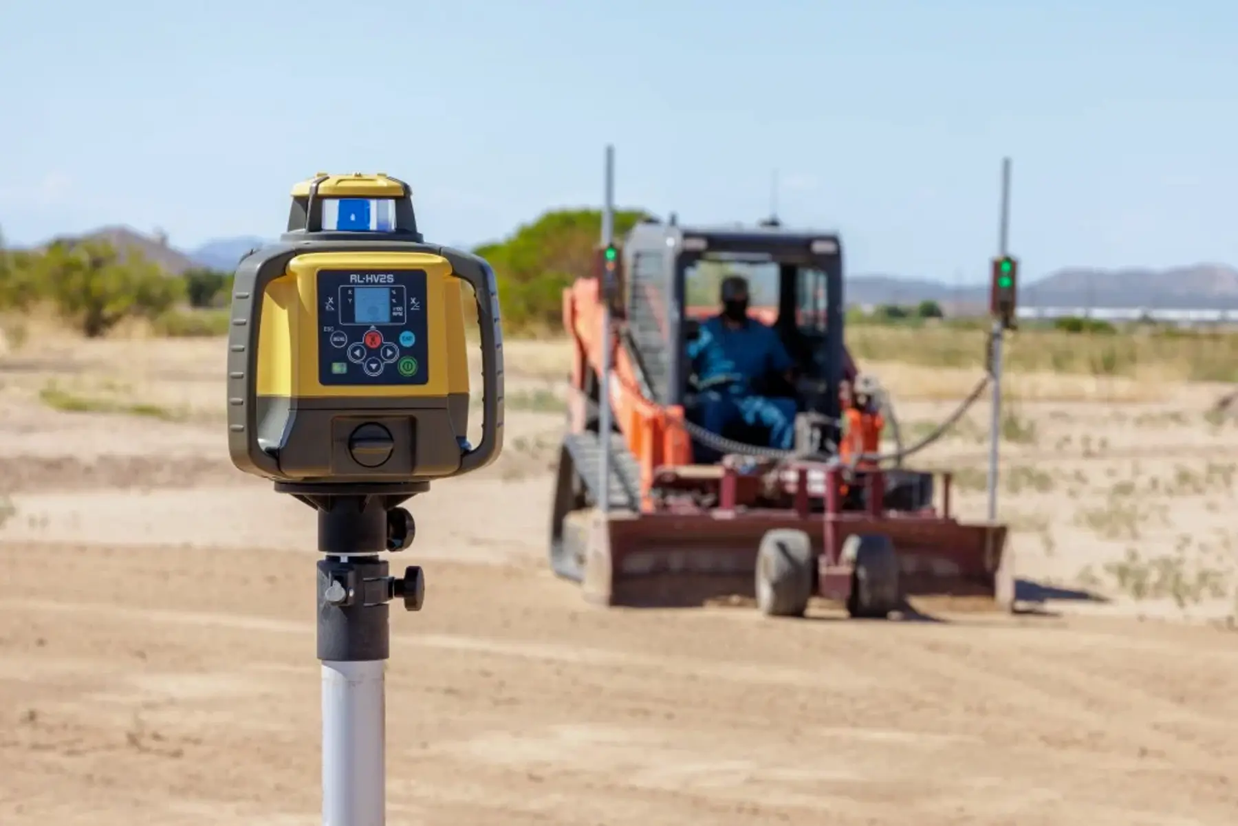A laser level stands in the foreground on a construction site, while a worker operates a small bulldozer in the background, grading the dirt. AdvanceTrac