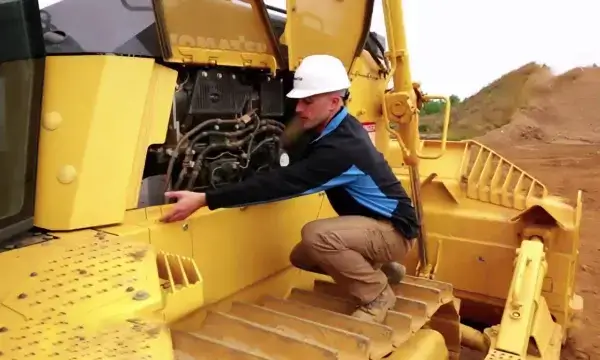 A person in a white hard hat and work clothes inspects the engine compartment of a yellow bulldozer at a construction site, with dirt and grassy areas in the background. AdvanceTrac