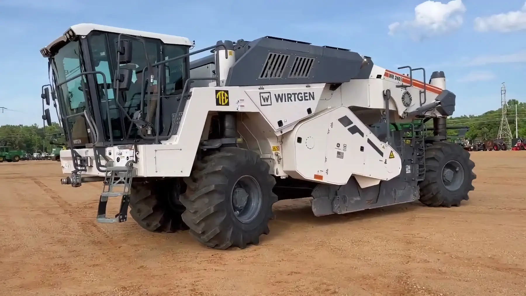 A large white Wirtgen industrial machine, known for its use as a reclaimer, with big black tires is parked on a dirt lot under a blue sky. AdvanceTrac