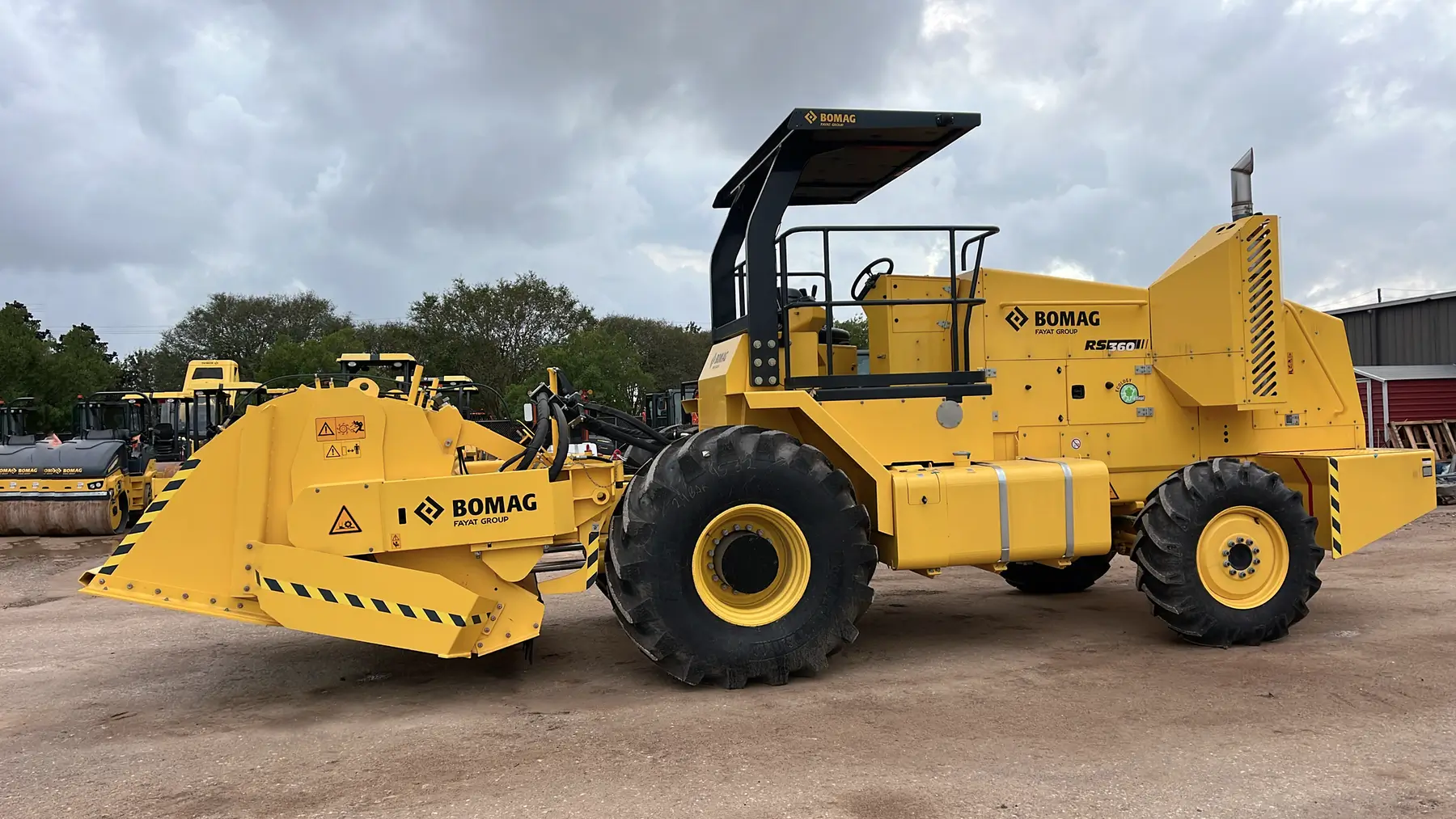 A large yellow BOMAG RS 250 road recycler machine is parked on a dirt lot with other equipment and trees in the background under a cloudy sky. AdvanceTrac