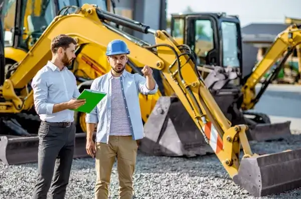 Two men standing in front of yellow excavators at a construction site; one wearing a blue hard hat and gesturing, the other holding a green folder and listening attentively. AdvanceTrac