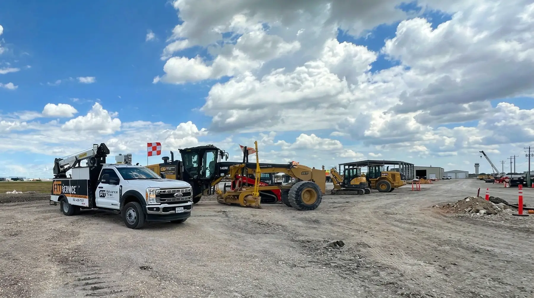 A construction site with trucks and heavy machinery on a dirt lot under a partly cloudy sky. Orange cones line the area, and a white utility truck is parked near the equipment. AdvanceTrac