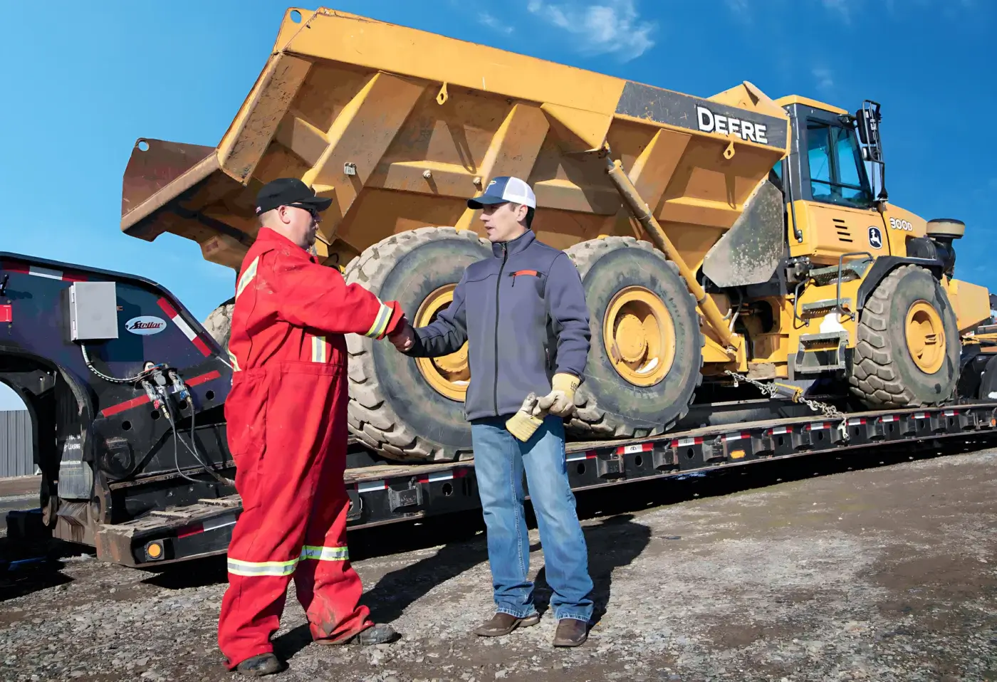 Two men, one in a red uniform and one in a jacket and jeans, shake hands in front of a large yellow construction vehicle loaded on a flatbed trailer under a blue sky. AdvanceTrac