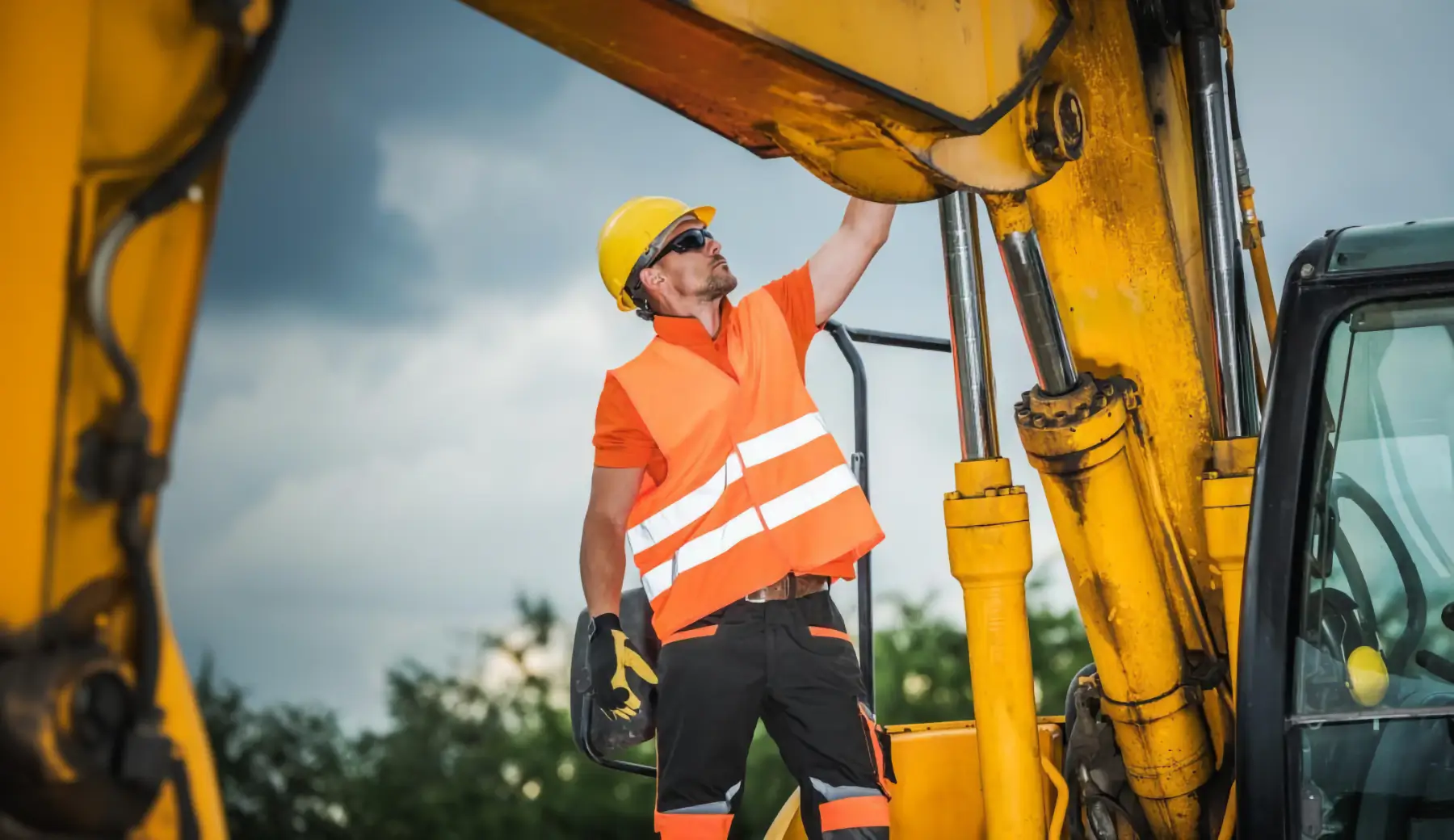 Construction worker in orange safety vest and yellow helmet inspects or repairs large yellow heavy machinery outdoors, with trees and cloudy sky in the background. AdvanceTrac