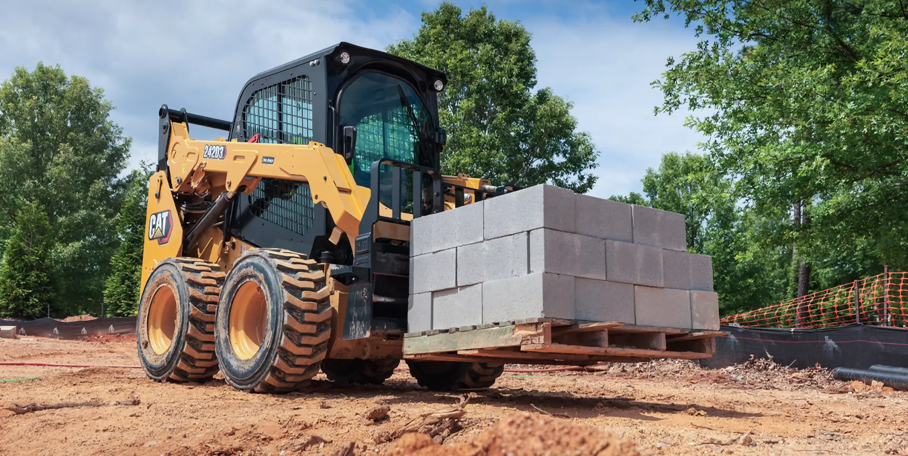 A yellow skid steer loader lifts a wooden pallet stacked with concrete blocks at a construction site with dirt ground and trees in the background. AdvanceTrac