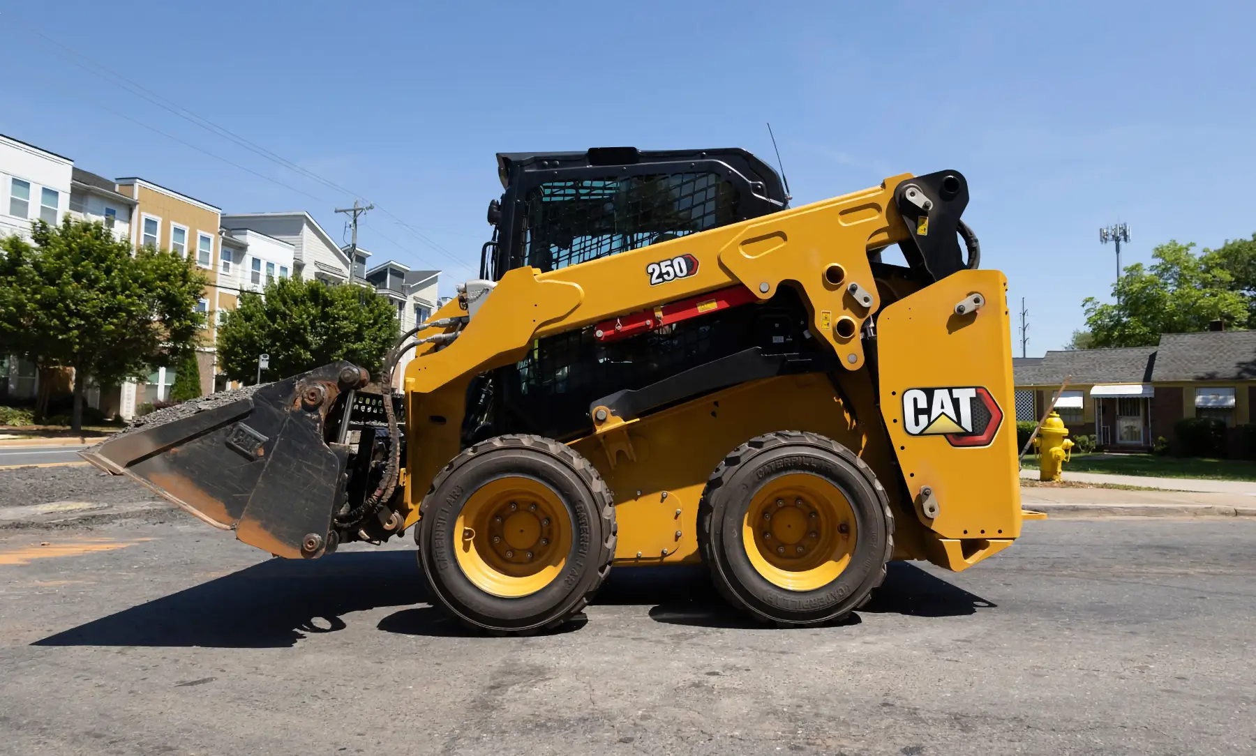 A yellow CAT skid steer loader parked on a paved street in front of residential buildings and trees, with its bucket attachment raised slightly off the ground. AdvanceTrac