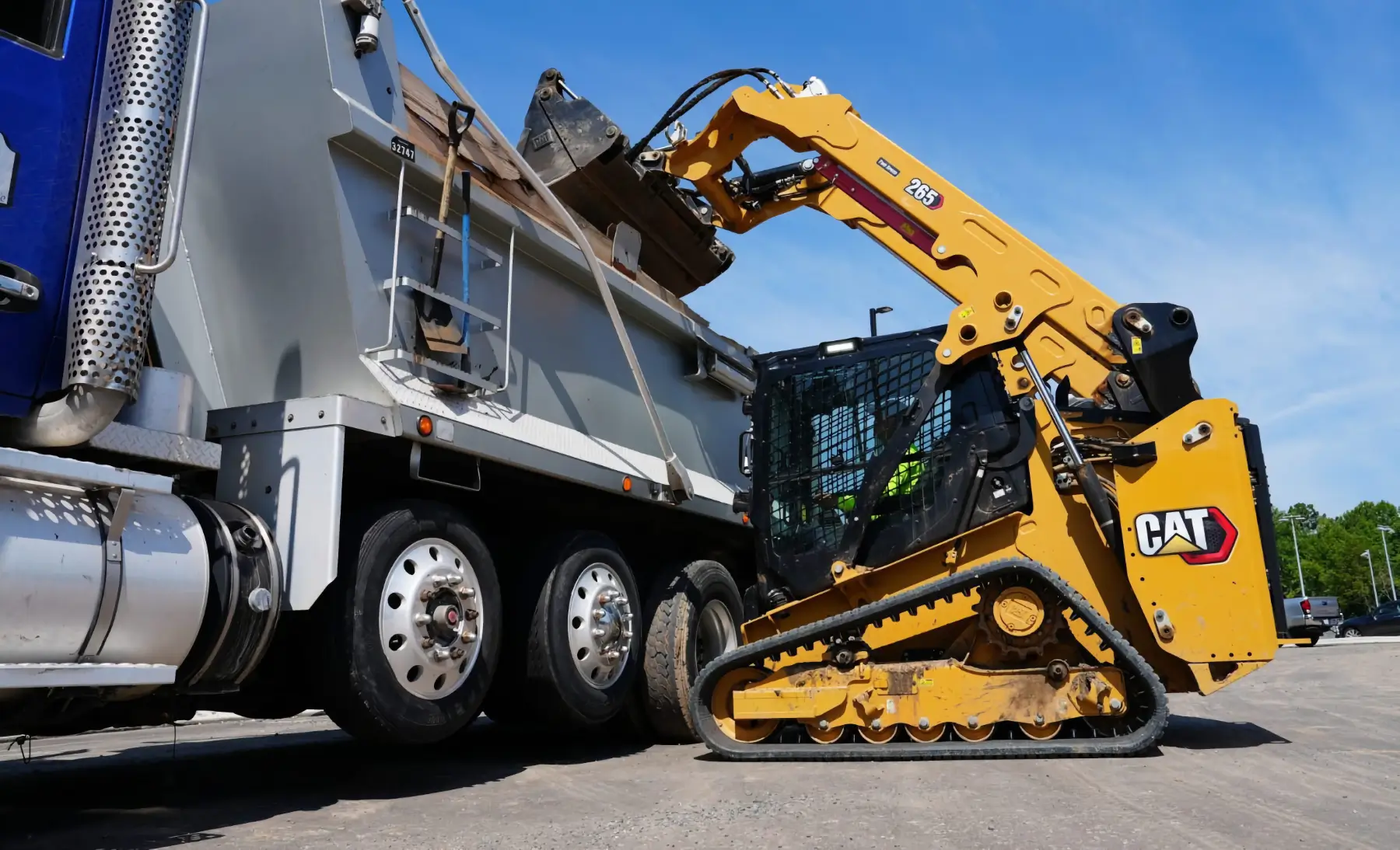 A yellow Caterpillar skid steer loader is lifting dirt into the bed of a large dump truck on a sunny day. Both vehicles, commonly used alongside track loaders for heavy-duty construction tasks, are on a paved surface, and trees are visible in the background. AdvanceTrac