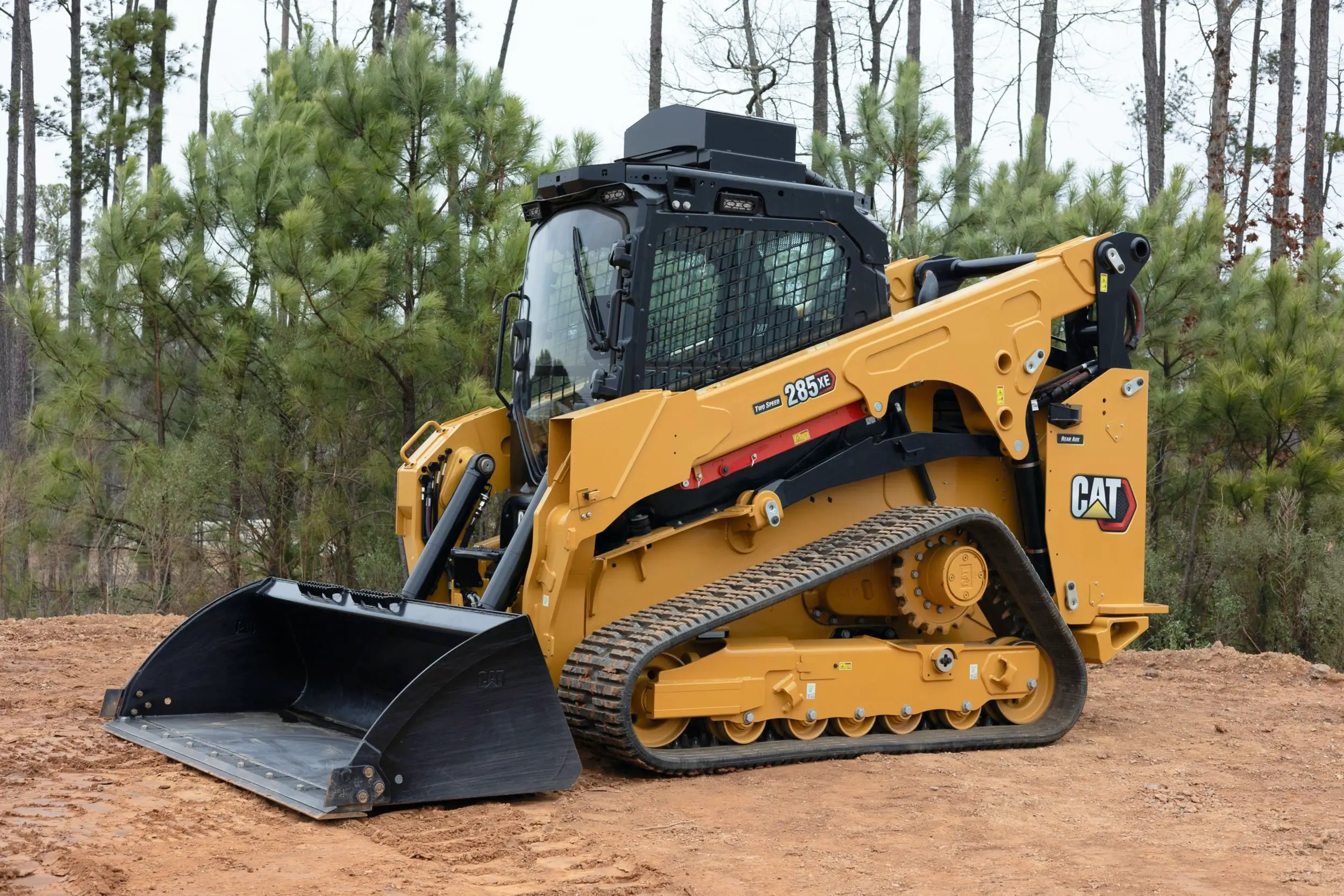 A yellow Caterpillar compact track loader with a front bucket attachment is parked on a dirt surface near pine trees, showcasing the versatility and efficiency of modern Track Loaders. AdvanceTrac