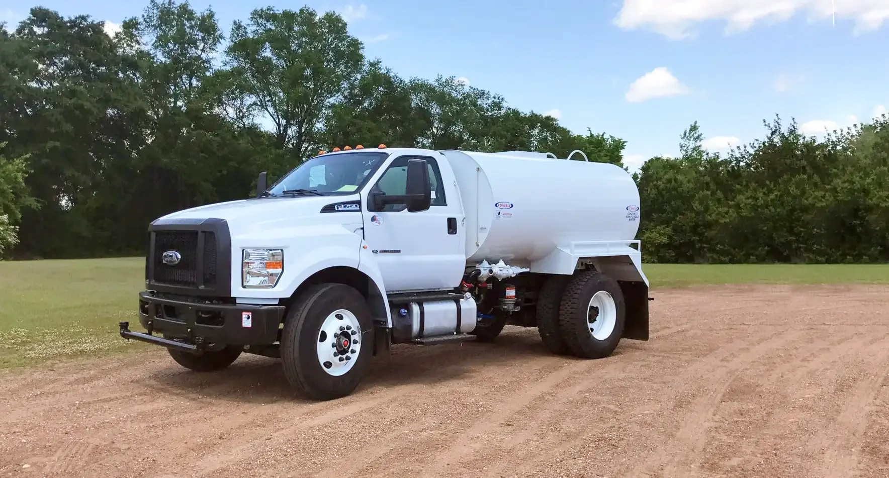 A white water tanker truck, equipped with essential water equipment, is parked on a dirt path with trees and grass in the background under a partly cloudy sky. AdvanceTrac