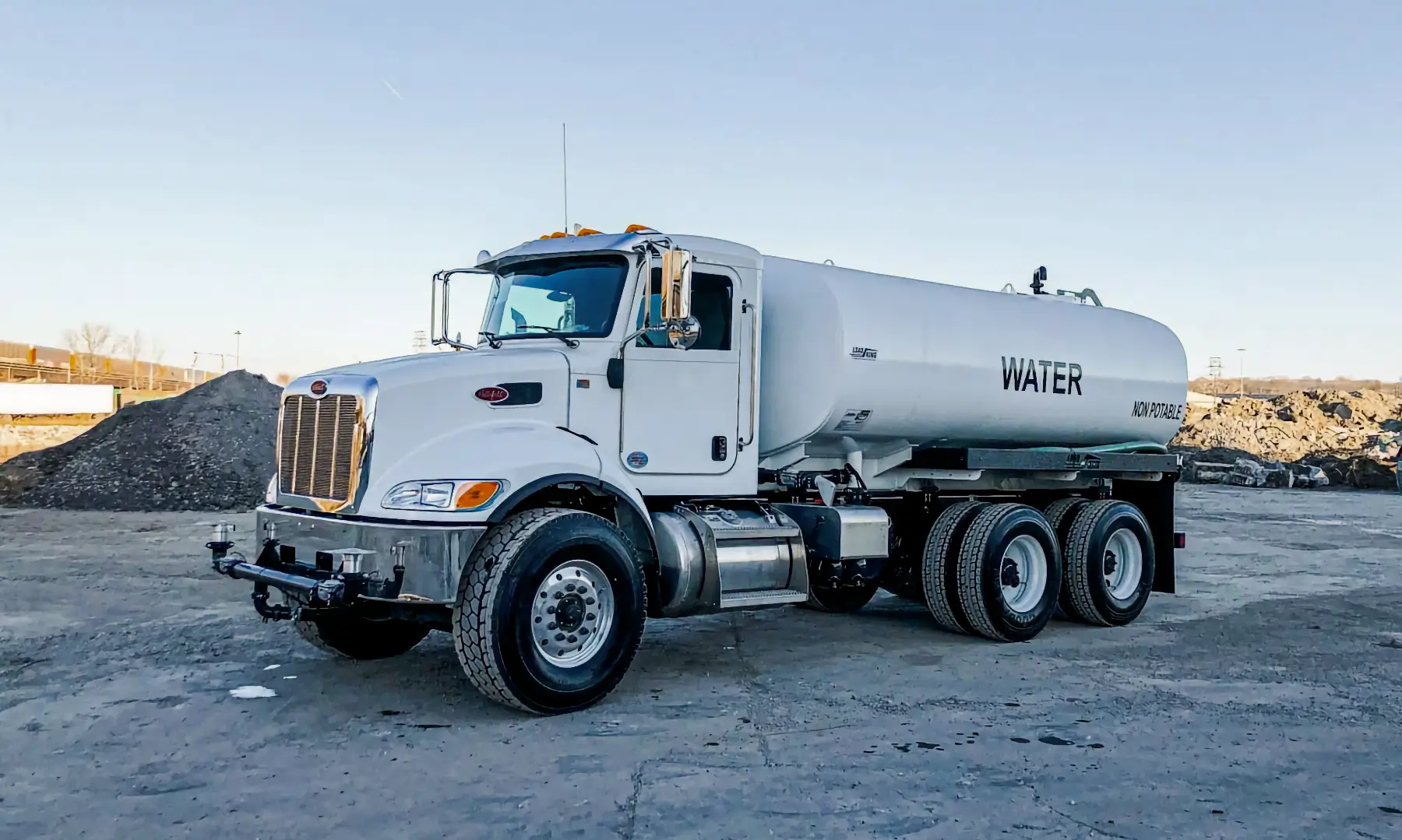 A white water tanker truck, part of essential water equipment, is parked on a gravel lot, with the word “WATER” printed in large letters on the side of the tank. The background includes piles of dirt and a clear sky. AdvanceTrac